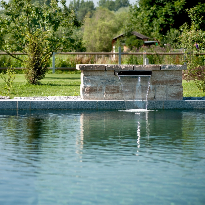 Eine Schwalldusche ist am Rand des Pools in eine GDM.KLASSIK wall-Mauer eingelassen und plätschert ins Wasser.